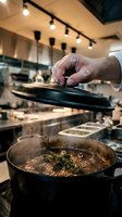 Professional chef covering cast iron skillet with lid while cooking aromatic herb-seasoned dish in modern commercial kitchen with track lighting.