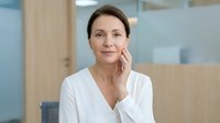 Middle-aged Caucasian woman touching face gently in modern office environment. Professional businesswoman skincare routine wellness concept.