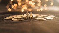 Miniature elderly couple figurines sitting on scattered coins with warm bokeh lights background representing retirement planning and financial security.