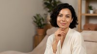 Middle-aged woman with dark wavy hair and olive complexion wearing cream cardigan, sitting on beige sofa in modern living room with potted plants and wooden shelves, portrait for lifestyle and wellness content.
