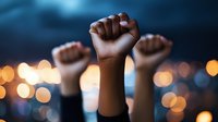 Raised fists silhouetted against blurred city lights at dusk symbolize unity, protest, and social movement. Dramatic bokeh effect creates powerful atmosphere.