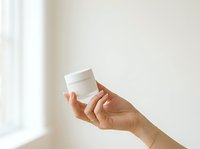 Unrecognizable person holding white cosmetic jar against neutral background with natural light, suitable for skincare and beauty product marketing.