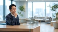 Thoughtful Asian male businessman in dark suit sitting at wooden desk with hand near face, contemplating in modern office with large windows and city view.