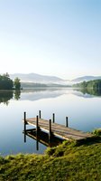 Wooden dock extending into calm lake with mountain reflections, surrounded by green grass and forested shoreline under clear sky for travel and nature concepts.