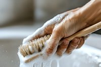 Close-up of hand scrubbing surface with wooden brush and soap foam, showing cleaning process in action. Natural bristles and bubbles create dynamic texture.