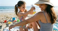 Young girl applying sunscreen to mother's shoulder during beach vacation, family sun protection and skincare routine on sandy shore with ocean background.