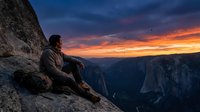 Man sitting on rocky cliff edge at sunset overlooking vast valley canyon landscape, contemplative solitude moment during golden hour, suitable for travel and lifestyle content.