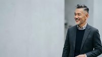 Mature Asian male professional in dark blazer and black sweater standing against neutral gray wall background, suitable for corporate, business, and leadership concepts.