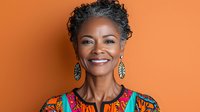 Confident mature African American woman with natural gray hair wearing colorful ethnic pattern dress, smiling warmly against orange background for wellness and lifestyle content.