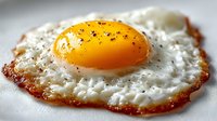 Perfectly cooked sunny-side up fried egg with golden yolk and crispy edges, seasoned with black pepper on white surface for breakfast and cooking concepts.