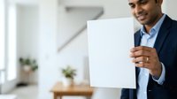 Young South Asian male professional in dark blazer and light blue shirt holding blank white card in modern office environment with blurred background and potted plants.