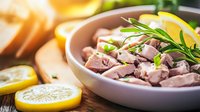 Fresh tuna chunks seasoned with herbs, lemon slices and rosemary in ceramic bowl, raw seafood preparation with citrus garnish on wooden surface.