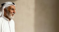 Elderly Middle Eastern man with gray beard wearing traditional white thobe and ghutra headwear, looking thoughtfully to the side against neutral beige background for cultural representation.