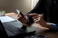Professional businessman using smartphone at modern office desk with laptop computer for mobile communication and digital workflow management.