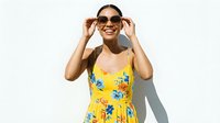 Young woman with dark hair and medium skin tone wearing yellow floral summer dress and sunglasses, smiling at camera against white background for fashion and lifestyle content.