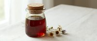 Glass jar with cork stopper filled with deep red herbal infusion or tincture, displayed on white linen surface with dried chamomile flowers nearby for natural wellness and alternative medicine.