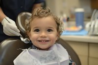 Smiling curly-haired toddler at dental appointment wearing protective bib while dentist examines teeth in modern medical office, promoting pediatric dental care and oral health.