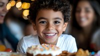 Young Latino boy with curly hair smiling brightly at birthday party with decorated cupcake and festive bokeh lights in background, capturing pure childhood joy.