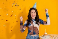 Young woman celebrating birthday with party hat and confetti on bright orange background, arms raised in joy with cake and candle nearby.
