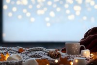 Cozy winter atmosphere with hot drink, warm knitted blanket and glowing fairy lights creating magical bokeh effect against soft blue background.