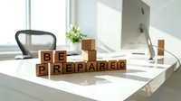 Wooden blocks spelling "BE PREPARED" arranged on a bright office desk with natural sunlight, featuring a computer monitor, chair, and potted plant, conveying readiness and business planning concepts.