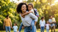 Happy African American mother carrying smiling daughter on back in sunny park during family gathering. Joyful parenting moments outdoors with warm sunlight.