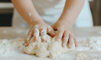 Hands kneading raw dough on floured wooden surface, close-up view showing flour-covered fingers pressing into soft, elastic bread dough during home baking process.