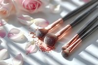 Professional makeup brushes with rose gold details on white surface with scattered pink rose petals and eyeshadow powder, natural window light creating soft shadows, beauty concept.