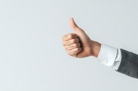 Male hand in business suit sleeve showing thumbs up gesture against light background, expressing approval and positive feedback in professional setting.