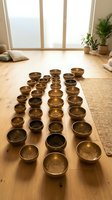 Multiple brass singing bowls arranged in rows on wooden floor in bright studio space with large windows, potted plants, and cushions, suitable for meditation, wellness, yoga instruction, and sound healing therapy content.