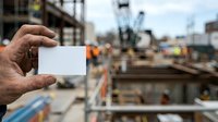 Close-up of male worker's hand holding blank white business card at active construction site with heavy machinery and building framework in background.