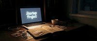 Dimly lit workspace with laptop displaying startup project text, open notebook, scattered photos, and coffee cup on wooden desk.