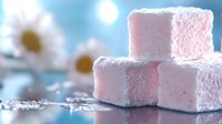 Pink marshmallow cubes dusted with powdered sugar on reflective surface against soft blue bokeh background with white flowers, sweet dessert confectionery closeup.