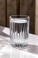 Crystal glass with pure water on marble surface casting dramatic shadows in natural light. Vertical ribbed pattern creates elegant reflections and refractions.