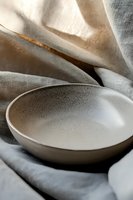 Minimalist ceramic bowl with speckled glaze texture on natural linen fabric background, creating soft shadows and folds in neutral beige and gray tones.