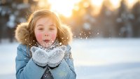 Young blonde girl in winter coat blowing snow from mittens during golden hour sunset. Magical winter moment with sparkling snowflakes in cold weather outdoors.