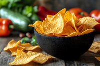 Crispy corn tortilla chips in black ceramic bowl on rustic wooden table with fresh vegetables in background, appetizing snack food composition.