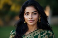 Young South Asian woman with long black hair wearing traditional green and gold embroidered dress and ornate gold earrings smiling at camera against blurred natural background.