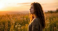 Young woman with curly hair in knit sweater enjoying peaceful sunset moment in wildflower meadow during golden hour for wellness and mindfulness content.