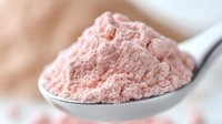 Pink protein powder supplement on metal spoon, macro view showing fine powder texture and soft pastel color against blurred background.