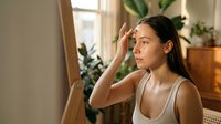 Young woman of East Asian descent applying skincare product to her face in a bright, plant-filled bedroom. Suitable for beauty, wellness, and skincare marketing.