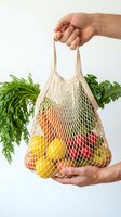 Hands holding a reusable cotton mesh bag filled with fresh fruits and vegetables on white background, eco-friendly shopping concept.
