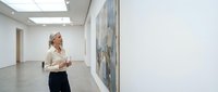 Professional mature woman with gray hair admiring contemporary artwork in modern gallery space with white walls and polished floors for cultural venues.