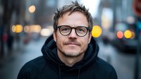 Caucasian man in black hoodie and glasses smiling at camera on blurred urban street background with bokeh lights. Close up portrait showing genuine happiness and confidence.