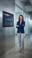 Professional businesswoman presenting data analytics dashboard in modern corporate office environment with arms crossed confidently.