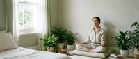 Woman in casual white shirt sitting cross-legged on bed in bright bedroom with potted plants, natural light from window, relaxed meditation or mindfulness practice setting.