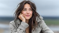 Young woman with long brown hair smiling gently while resting chin on hand, wearing cozy gray hoodie against blurred natural landscape background.