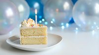 Vanilla birthday cake slice with lit candle and cream frosting on white plate against festive balloon background for celebration occasions.