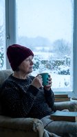 Senior Caucasian woman in burgundy knit hat enjoying hot beverage by snowy window on cozy winter morning at home.