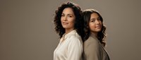 Two confident Latina women standing back to back against a neutral background, showcasing strength, bond, and elegance in casual attire.